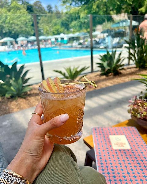 A hand holds a drink with a garnish near a pool area. There are lounge chairs and people by the pool, with clear, sunny weather.