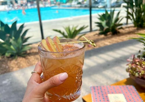 A hand holds a drink with a garnish near a pool area. There are lounge chairs and people by the pool, with clear, sunny weather.