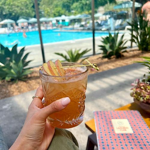 A hand holds a drink with a garnish near a pool area. There are lounge chairs and people by the pool, with clear, sunny weather.