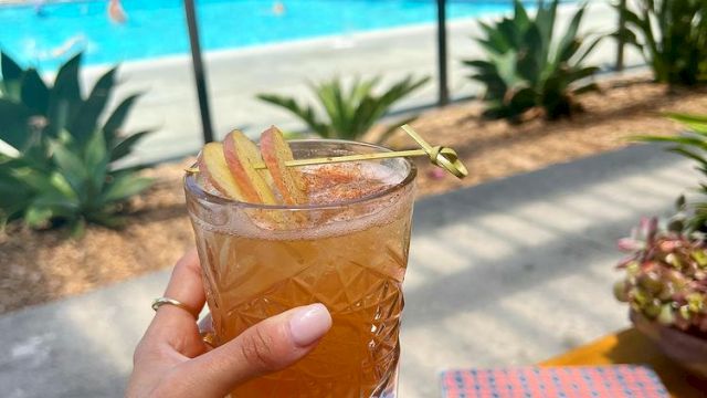 A hand holds a drink with a garnish near a pool area. There are lounge chairs and people by the pool, with clear, sunny weather.