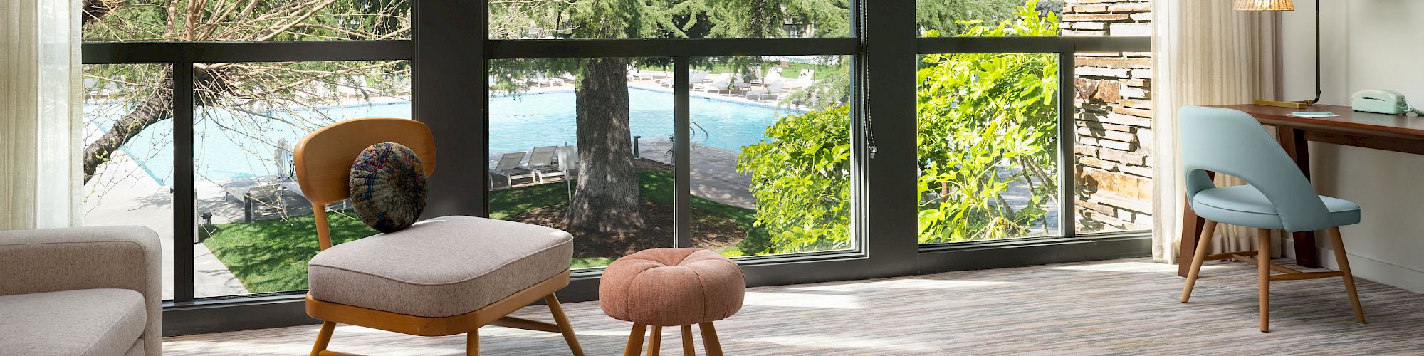 A modern living room with large windows overlooking a lush outdoor scene, featuring comfortable seating and a workspace near the window.