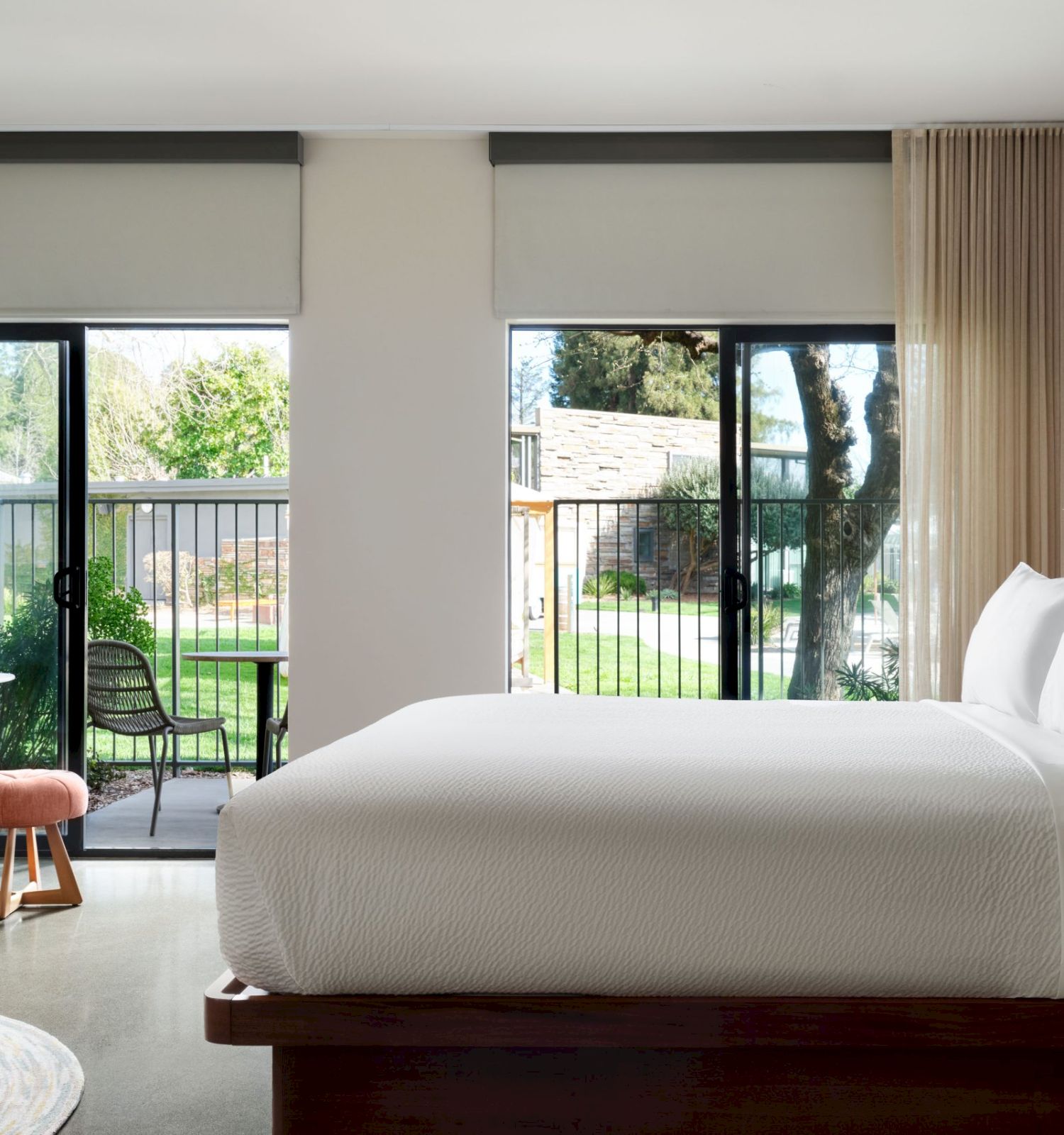 A modern bedroom with a large bed, two windows with sheer curtains, a small desk, a chair, and a view of a balcony with outdoor greenery visible.