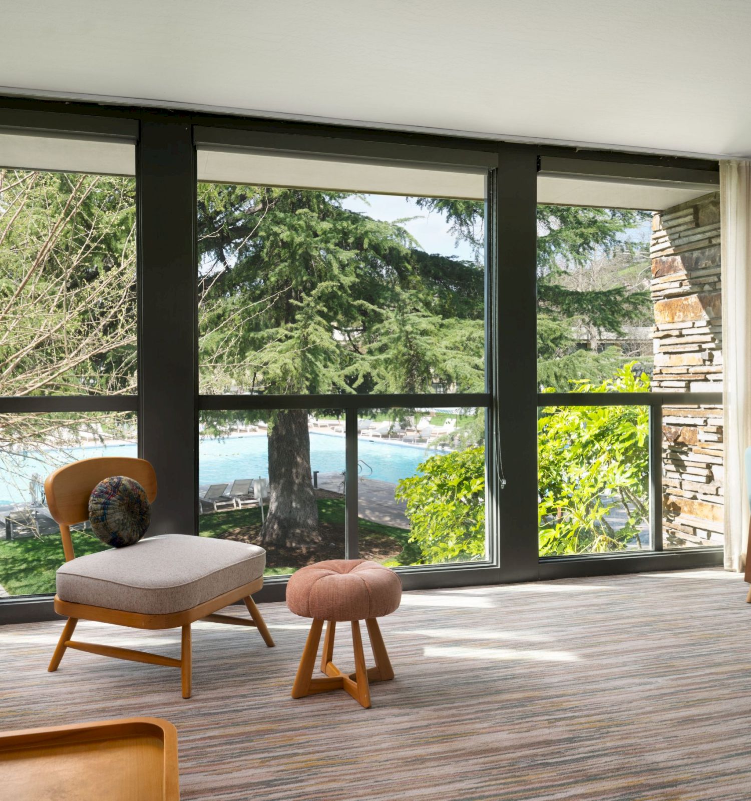 A modern living room features large windows showing a scenic outdoor view, with minimalist furniture, a desk, and a wall-mounted TV.