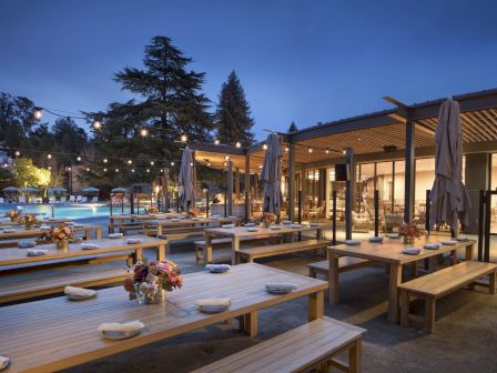 An outdoor dining area with wooden benches and tables set for guests, adorned with string lights and flower arrangements, captured at twilight.