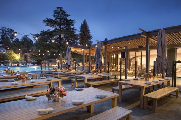 An outdoor dining area with wooden benches and tables set for guests, adorned with string lights and flower arrangements, captured at twilight.