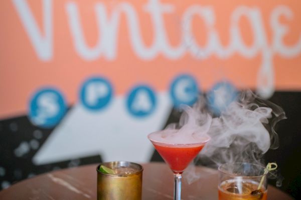 The image shows three cocktails on a table with a vintage-themed background displaying the word 