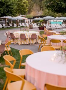 The image shows an outdoor event setup with round tables covered in white cloth and yellow chairs, situated near a pool area with sun loungers and umbrellas.