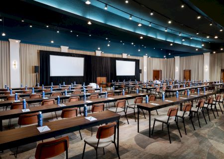 This image shows a large conference room set up with rows of tables and chairs, two large screens at the front, and water bottles on each table.