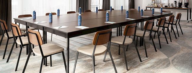 A conference room with a large, rectangular table surrounded by chairs; water bottles and notepads are on the table, and there's a refreshment station.