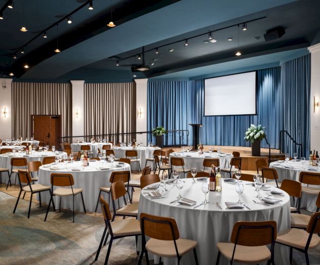 The image shows a spacious, elegant banquet hall set up for an event, with round tables covered in white tablecloths, chairs, and a stage with a podium.