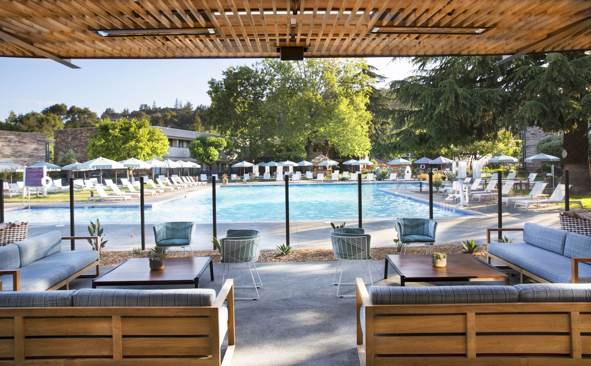 A view of a swimming pool from an outdoor seating area with sofas and chairs, surrounded by trees and buildings in the background.