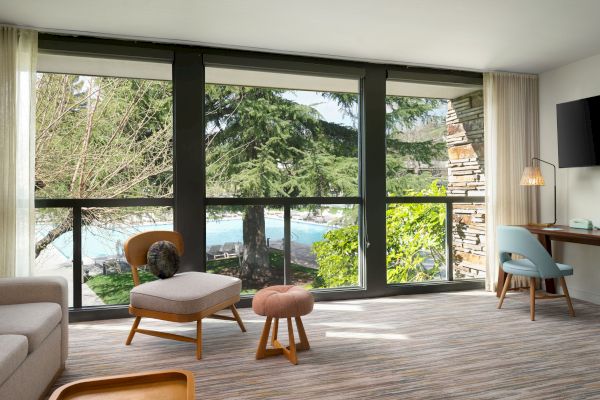 A modern living room features large windows with a nature view, contemporary furniture, a desk, a TV, and a beige carpet and walls.