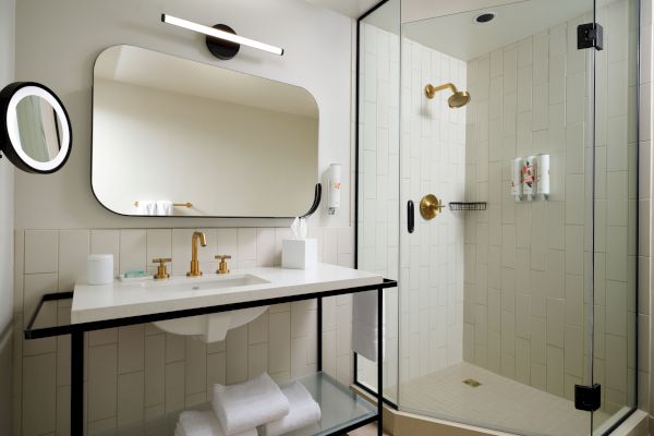 A modern bathroom with a glass-enclosed shower, white sink counter, gold fixtures, large mirror, and neatly folded towels on a shelf underneath.