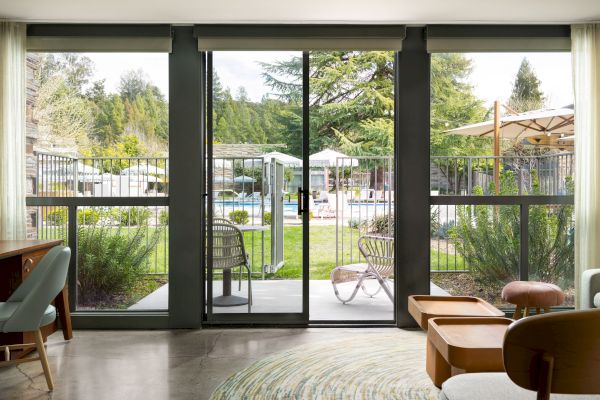 This image features a view from a living area through large glass doors, overlooking a garden with patio furniture and a pool surrounded by greenery.