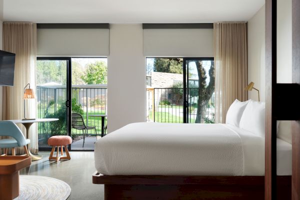 A modern hotel room with a large bed, a desk, chairs, and large windows leading to a balcony with a view of the outdoor area.