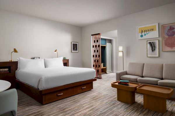 This image shows a modern hotel room with a bed, sofa, coffee table, wall art, desk, and a sliding wooden door separating the space.