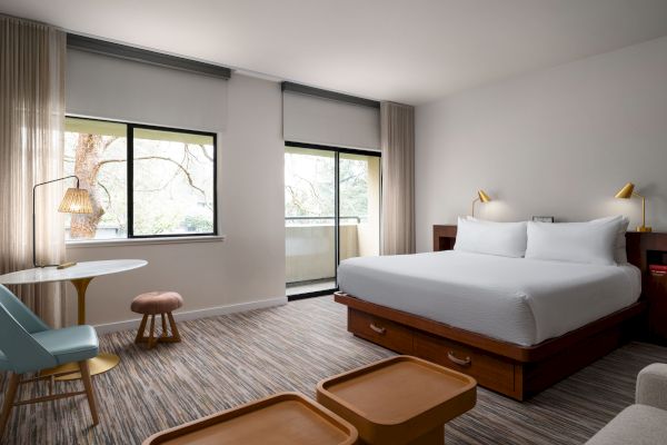 A cozy, modern hotel room with a king-sized bed, two large windows, a small table with a chair and stool, and two wooden coffee tables.