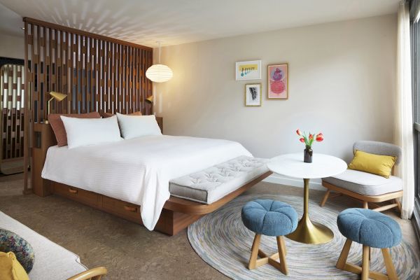 This image shows a modern, minimalist bedroom with a large bed, round table, blue stools, chair, and wall decor. Natural light fills the room.