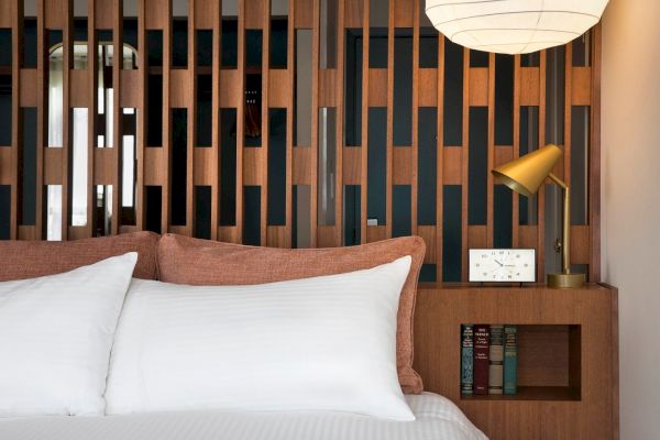 A cozy bedroom with a white bed, wooden headboard, books, a small white clock, and a golden lamp underneath a hanging paper lantern.