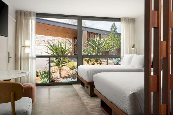 A modern hotel room with two beds, a large window overlooking greenery, a small round table with chairs, and a flat-screen TV on the wall.