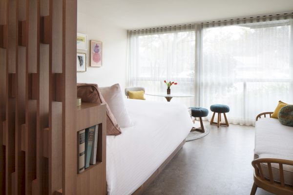 The image shows a modern, well-lit room with a bed, a small table with tulips, and stools by a large window with sheer curtains.