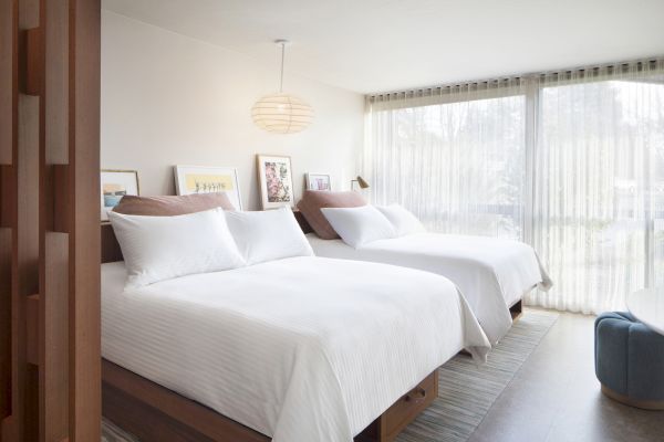 The image shows a modern bedroom with two double beds, white bedding, a hanging light, artwork, large windows with sheer curtains, and a seating area.