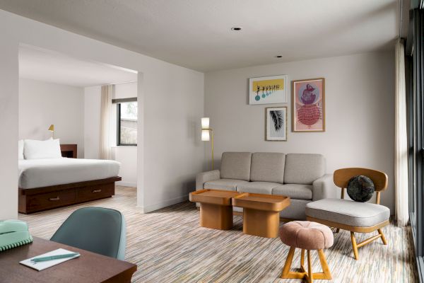 A modern hotel room features a living area with a sofa, coffee tables, chairs, and wall art, next to a bedroom with a double bed and a window.