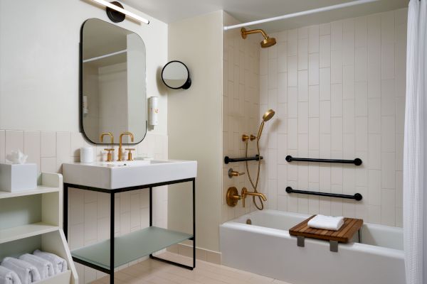 A modern bathroom with a black-framed mirror, sink, and shelving, featuring golden fixtures in the shower, and a wooden bath tray on the tub.