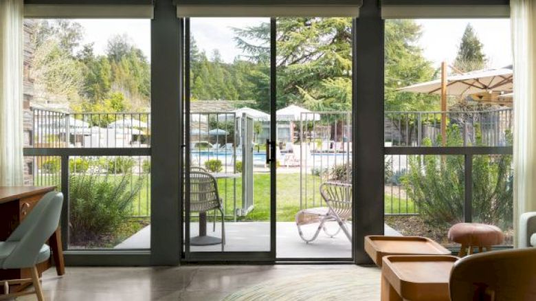 The image shows a modern living room that opens up to a patio with large glass doors, offering a view of an outdoor pool and seating area.