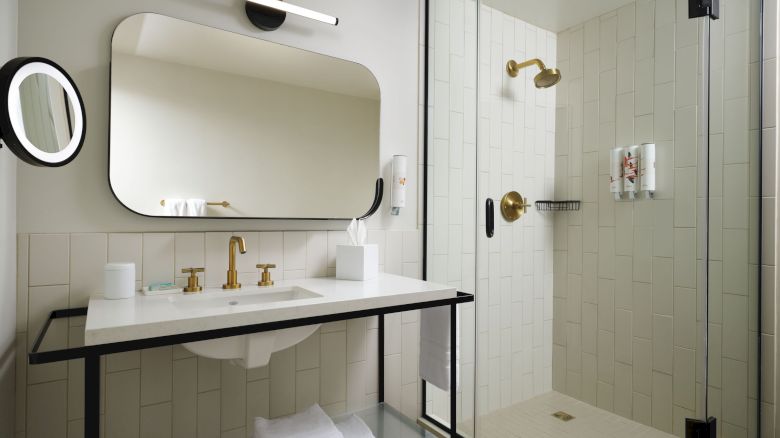 A modern bathroom with a vanity, large mirror, and glass shower enclosure with gold fixtures. Neat and minimalist design with white and black accents.