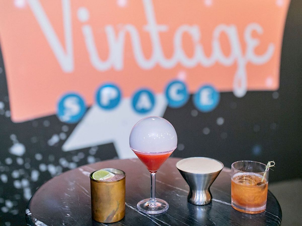 A table with four unique drinks against a "Vintage Space" sign background featuring cocktails in various glasses.