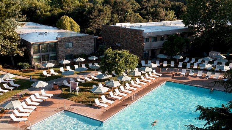 This image shows an outdoor swimming pool area with lounge chairs and umbrellas, surrounded by trees and buildings in the background.