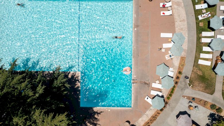 An aerial view of a swimming pool surrounded by lounge chairs and umbrellas, with a few people swimming and relaxing in the water.