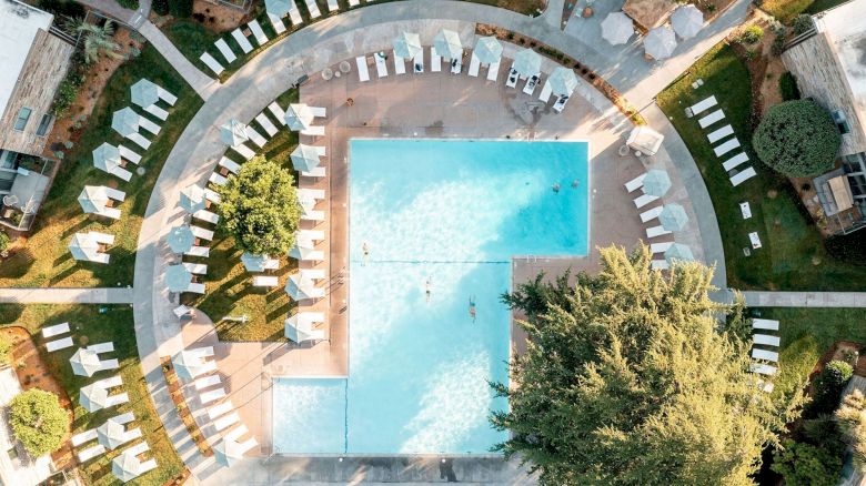 The image shows an aerial view of a large rectangular swimming pool surrounded by numerous lounge chairs and trees, within a landscaped area.