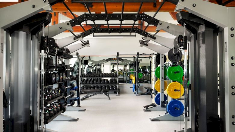 The image shows a well-equipped gym with various exercise machines, weights, and colorful weight plates set up in an open layout.