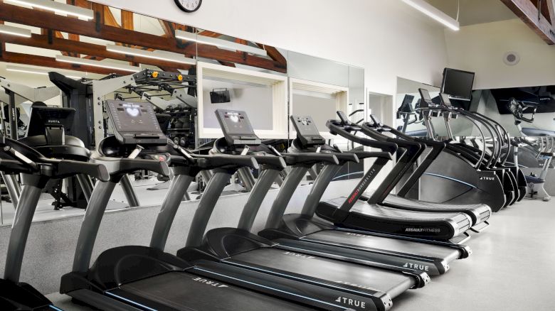 The image shows a gym with several treadmills lined up in front of large mirrors and additional workout equipment in the background.