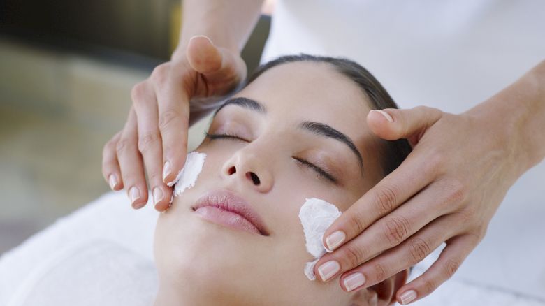 A person is receiving a facial treatment, with a professional applying cream to their face while they relax on a spa bed.