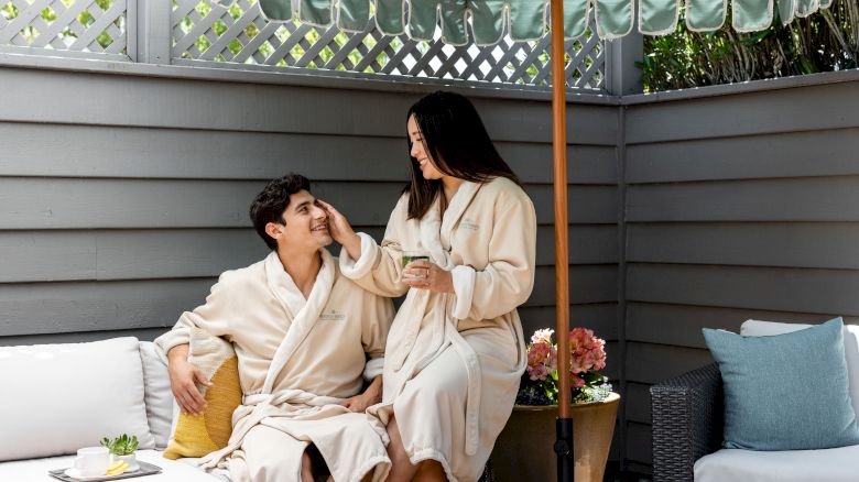 A couple in bathrobes sits on a patio, smiling and enjoying a relaxed moment under a sunshade. Comfortable seating and plants are visible.