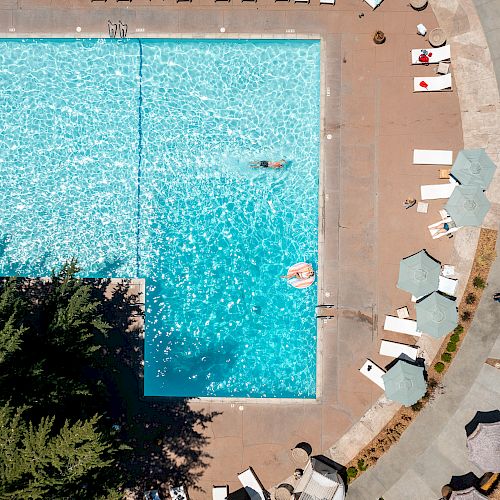 An aerial view of a swimming pool with people swimming, surrounded by lounge chairs, umbrellas, and a paved walkway in a lush area with trees.