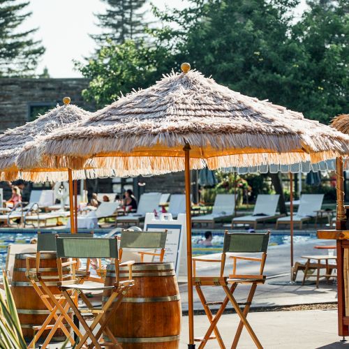 The image shows an outdoor seating area with wooden stools and tables under straw parasols near a pool, surrounded by greenery and lounge chairs.