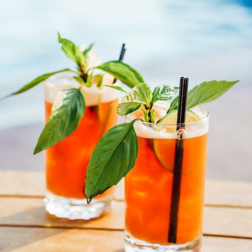 Two glasses of orange-colored drinks with ice, garnished with fresh green leaves and straws, placed on a wooden surface near a pool.