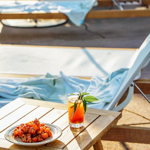 A lounge chair with a blue towel beside a table holding a glass of juice and a bowl of food is shown in a sunlit outdoor setting.