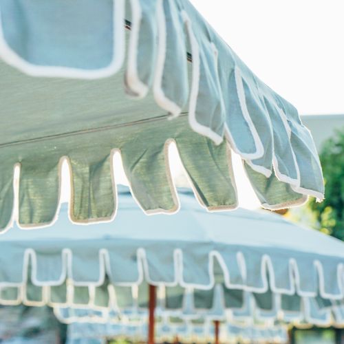 The image shows a close-up view of a light blue patio umbrella with white fringe detailing at the edge, set outdoors in a sunny environment.