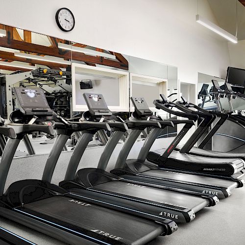 The image shows a gym with several treadmills and other fitness equipment, with a wall-mounted clock and mirrors.