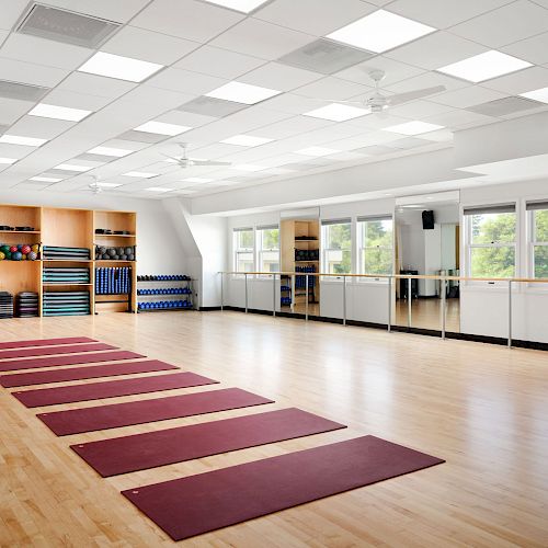 A spacious, well-lit yoga studio with red mats laid out on the wooden floor, large mirrors, windows, and shelves with yoga equipment in the background.