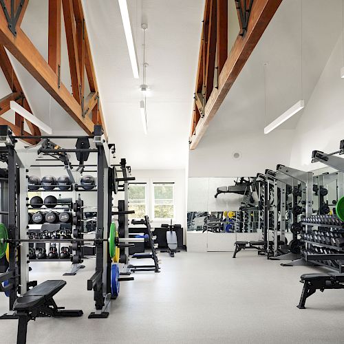 The image shows a well-equipped gym with free weights, weight machines, benches, and mirrors, featuring high wooden ceilings and ample lighting.