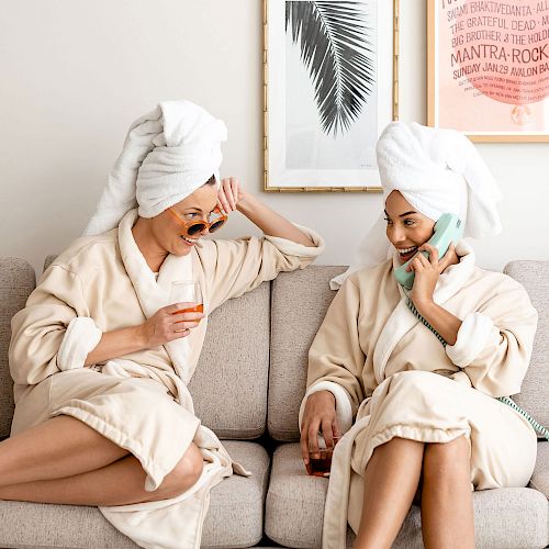 Two people in bathrobes and towels on their heads are sitting on a couch, laughing, one wearing sunglasses, and the other talking on a phone and holding a drink.