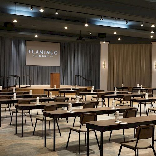 A conference room setup with desks and chairs facing a stage with a podium and screen labeled 