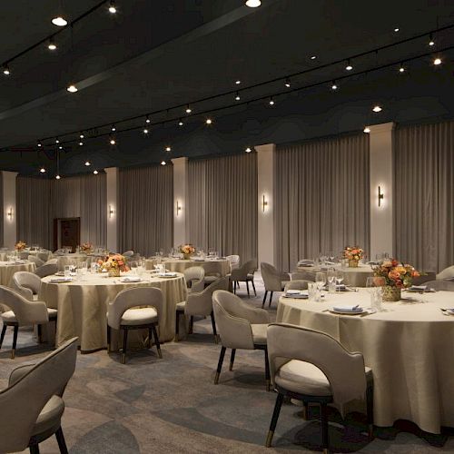 A banquet hall with round tables set with linens, flowers, and tableware. Several cushioned chairs surround each table, with curtains in the background.