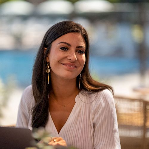 A woman is sitting outdoors at a table with a laptop, smiling and looking off to the side.